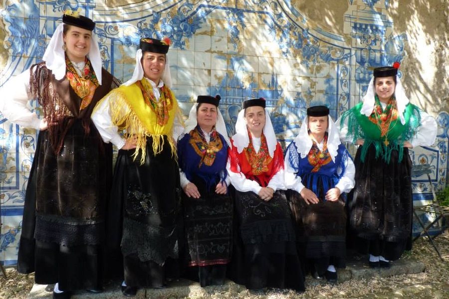 Elementos do grupo de folclore com trajes tradicionais, lenços brancos e xailes coloridos, diante de um painel de azulejos azuis