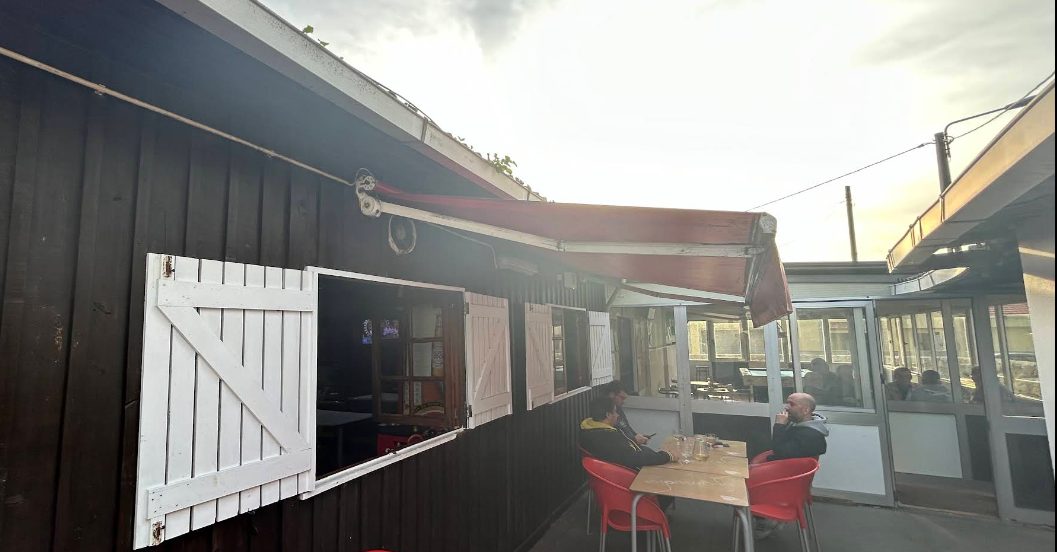 Esplanada exterior do Bar do Clube Académico, com toldo, mesas e a casa de madeira do clube