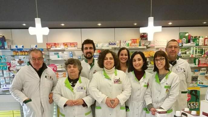 A equipa da Farmácia Gramaxo, de bata branca, ao balcão da farmácia, com as prateleiras e as cruzes verdes em fundo