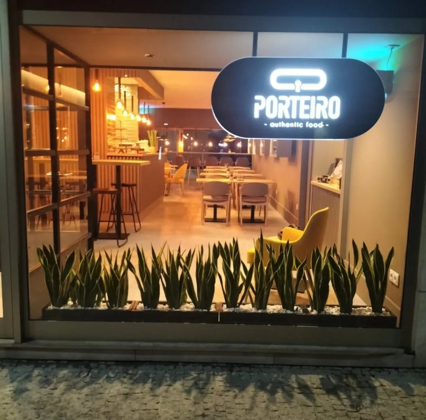 Montra do Porteiro, em Pedras Rubras, ao anoitecer, com o letreiro iluminado e a sala de madeira e luz quente lá dentro