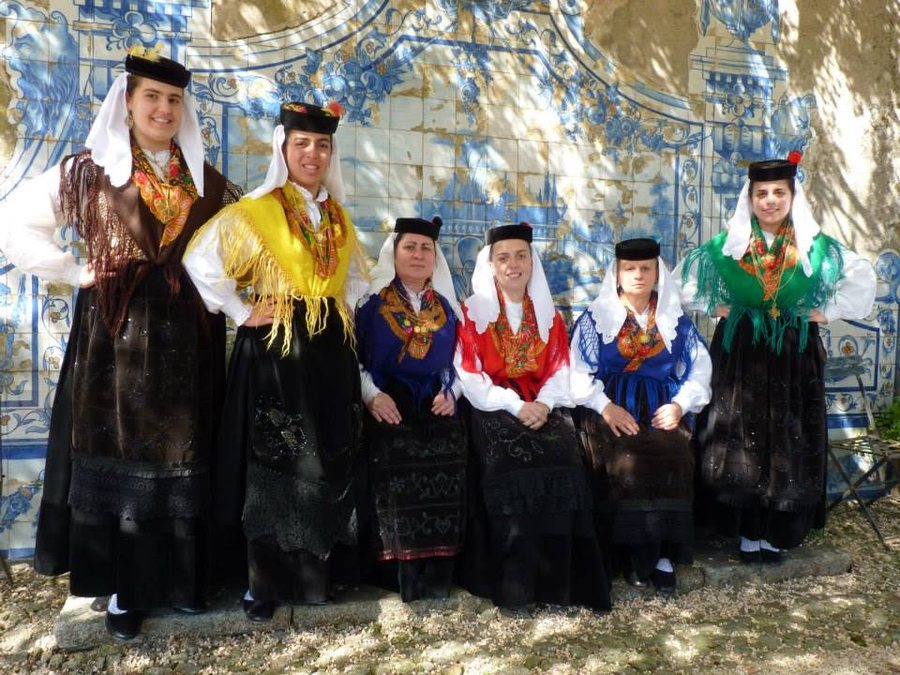 Seis elementos do grupo de folclore com trajes tradicionais — lenços brancos, chapéus pretos e xailes coloridos — encostados a um painel de azulejos azuis
