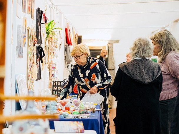 Visitantes a ver peças de artesanato e bijuteria expostas em mesas e nas paredes brancas do Celeiro dos Cónegos